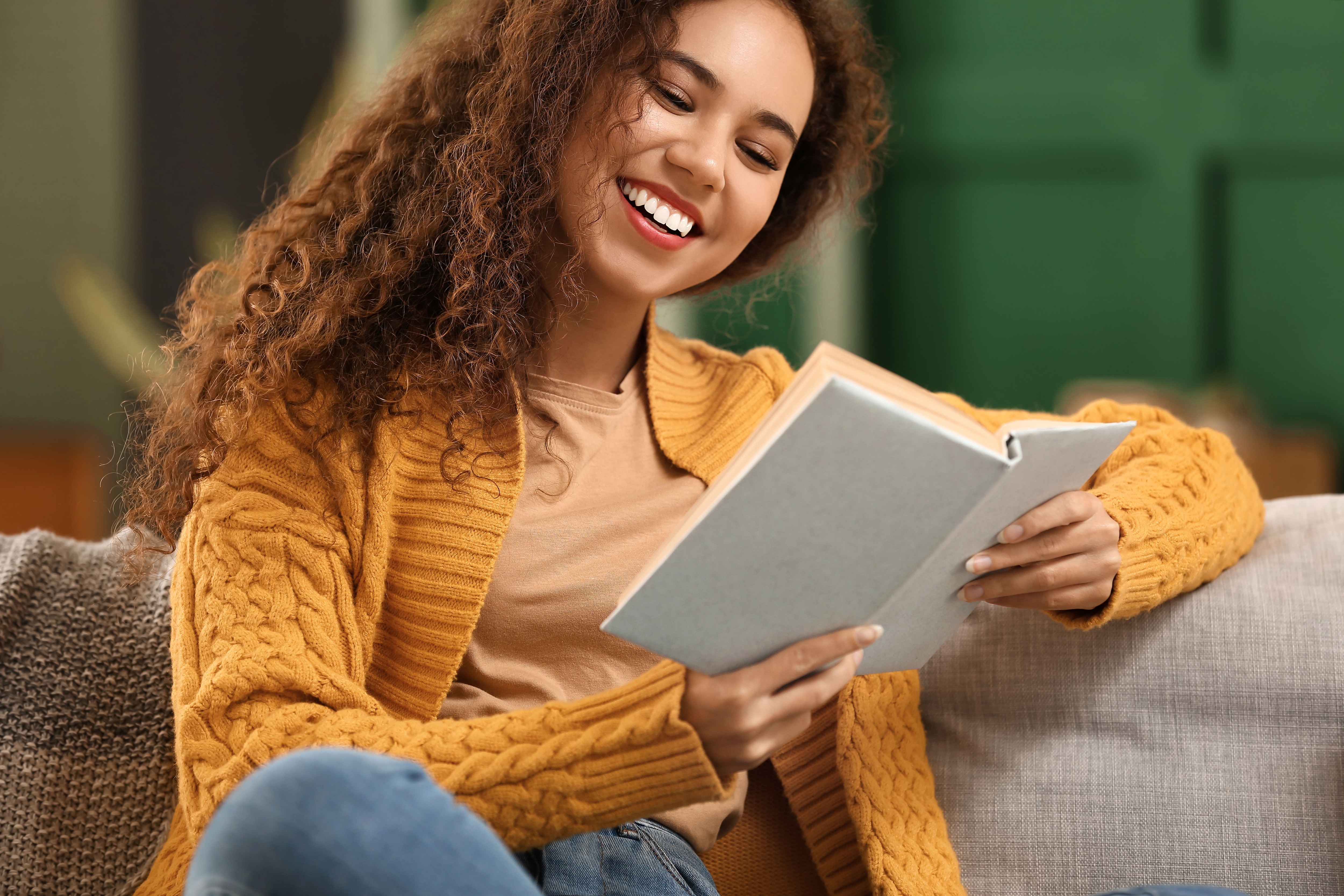 Smiling woman reading a book on a couch while wearing a mustard yellow knit sweater.jpeg