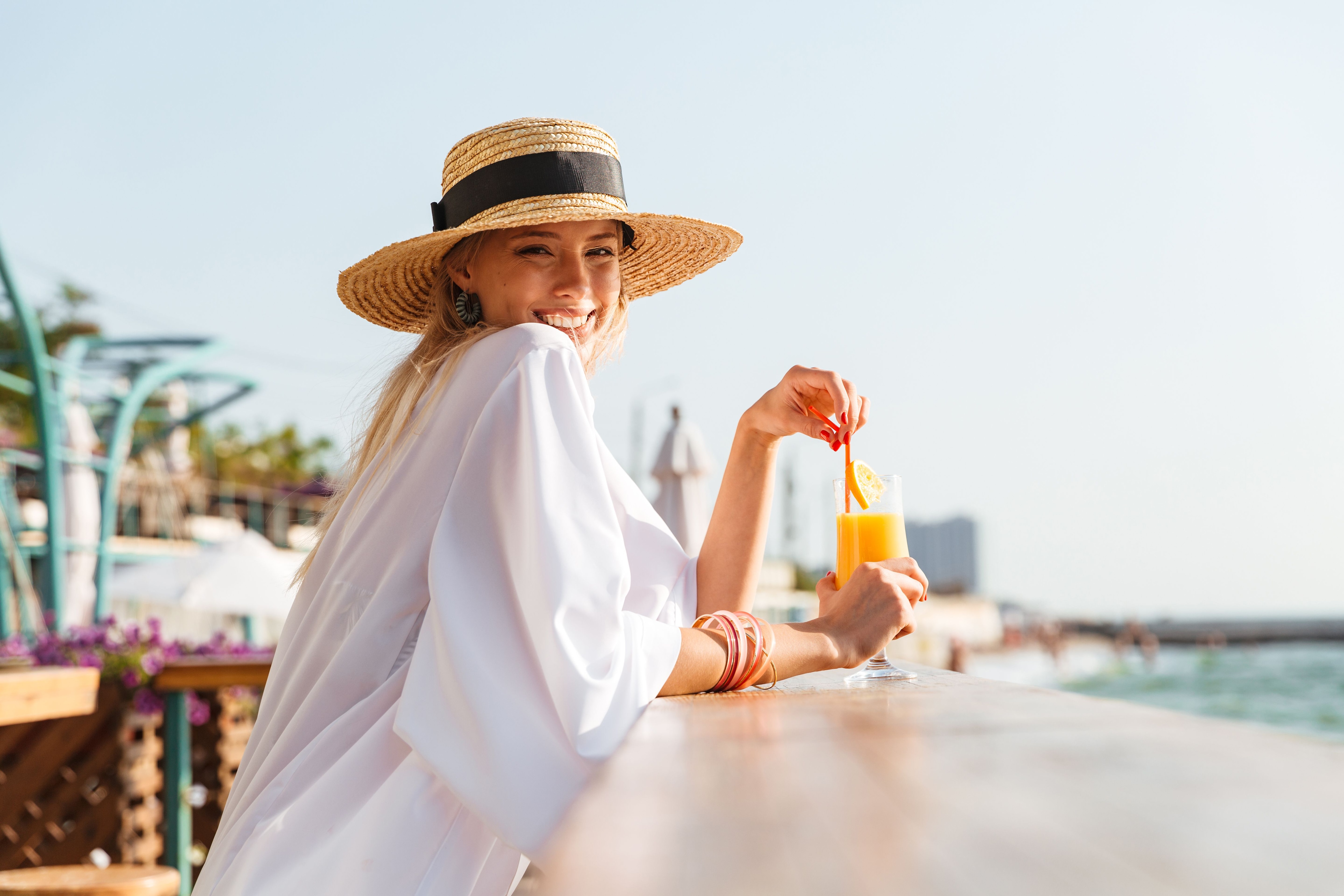 A smiling woman in a straw hat sits at a seaside bar on a beach vacation.jpeg
