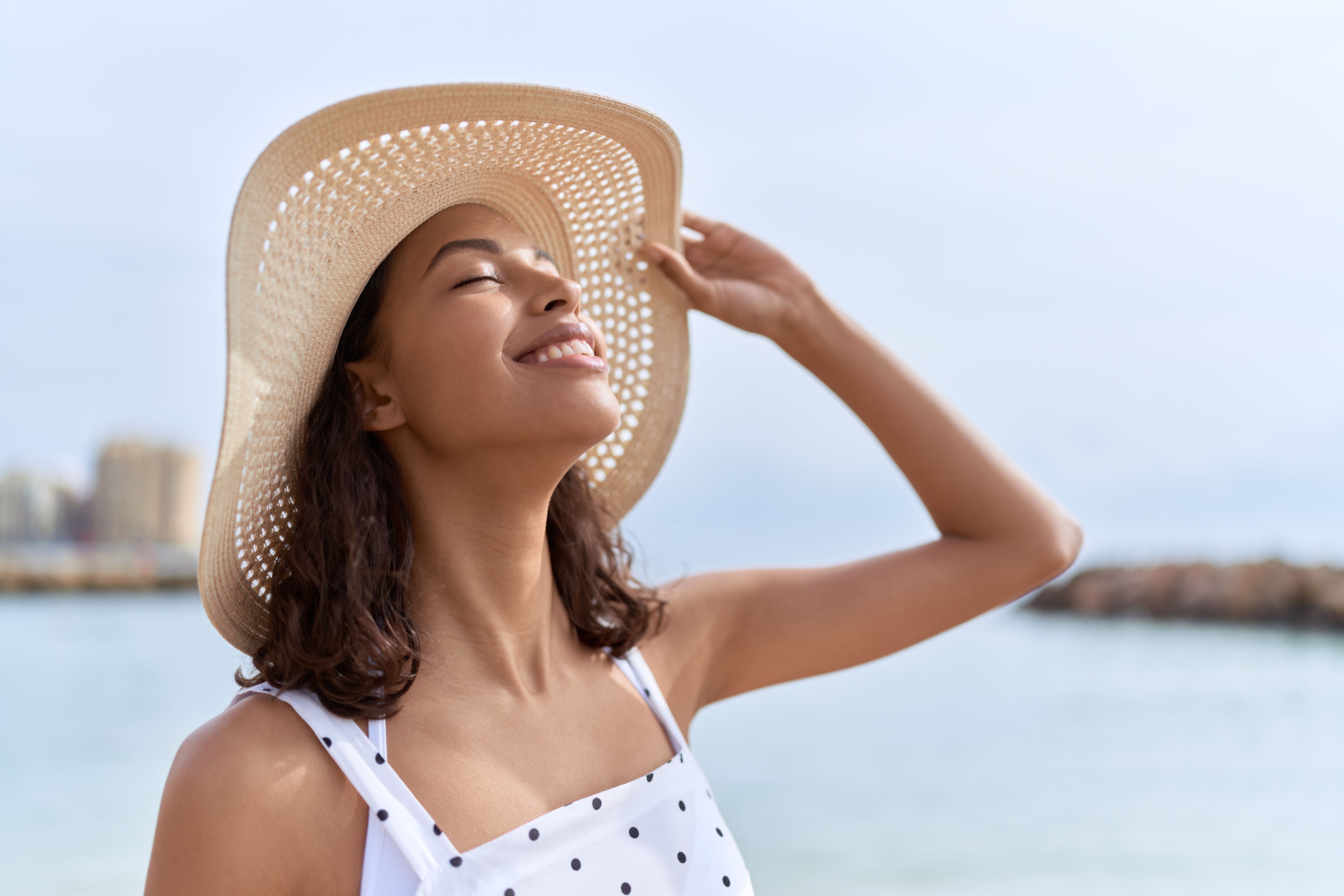 A smiling woman with closed eyes wears a polka dot dress and woven hat during summer.jpeg